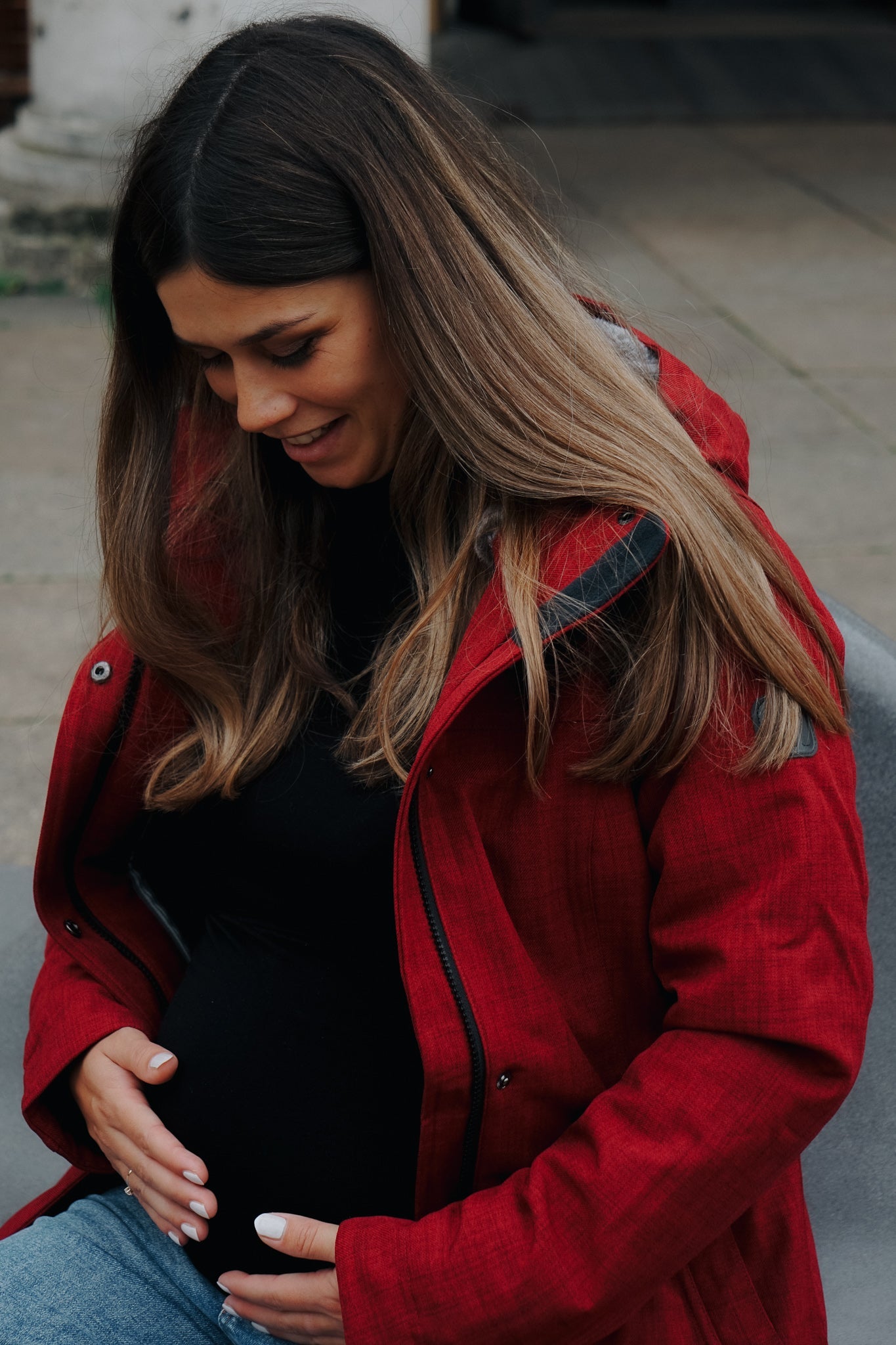 Schwangere Frau mit langen Haaren und dunkelroter Tragejacke haelt Babybauch