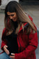 Schwangere Frau mit langen Haaren und dunkelroter Tragejacke haelt Babybauch